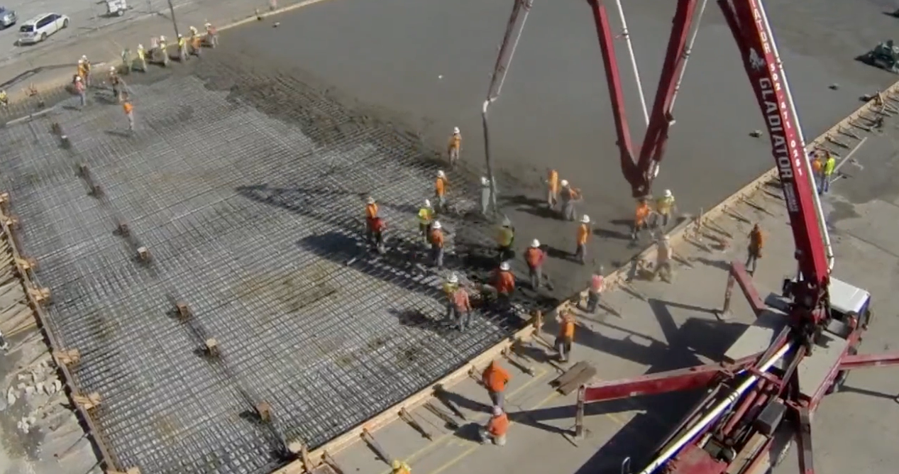 Construction workers pouring concrete on a large rebar slab beside a red pump truck boom