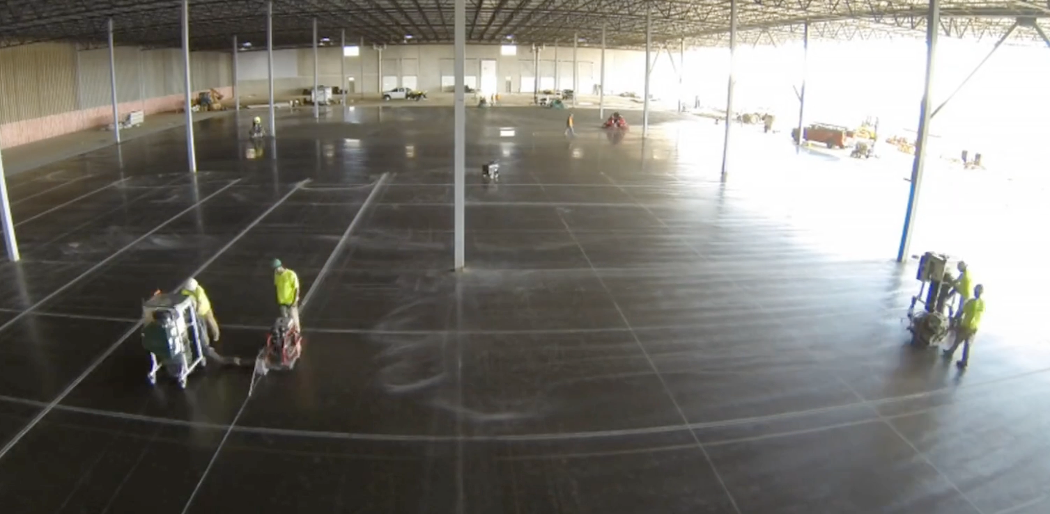 Workers in high-visibility vests inside a large empty warehouse, cleaning or marking the floor.