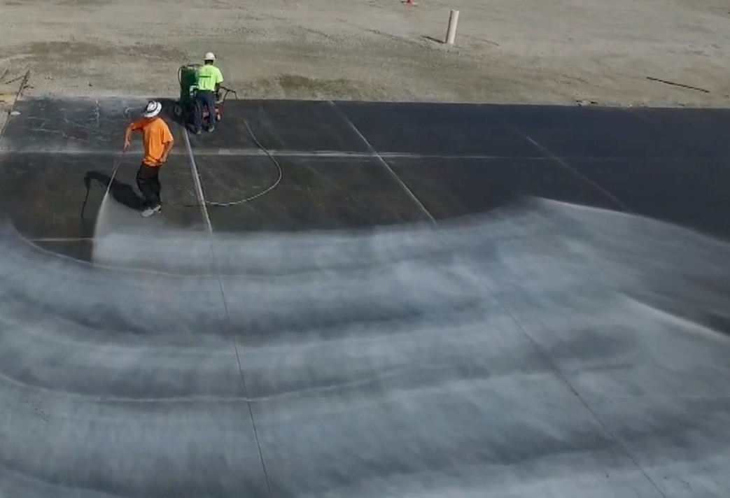Two workers in orange and green gear spraying water on a concrete surface at a worksite