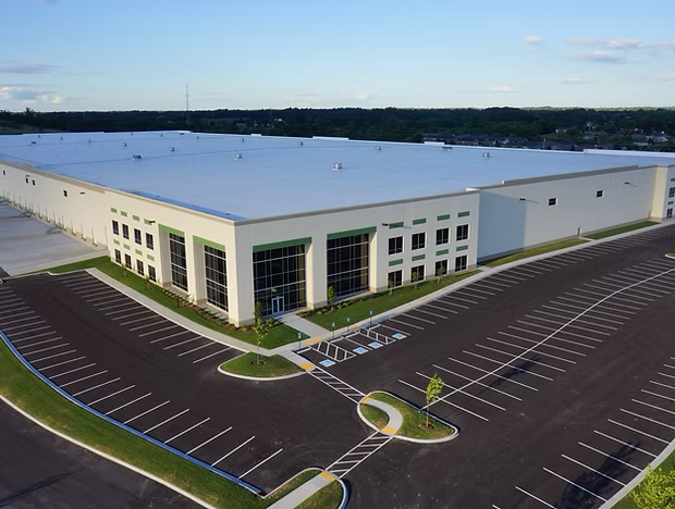 Aerial view of a large white commercial building with an empty parking lot and surrounding trees.