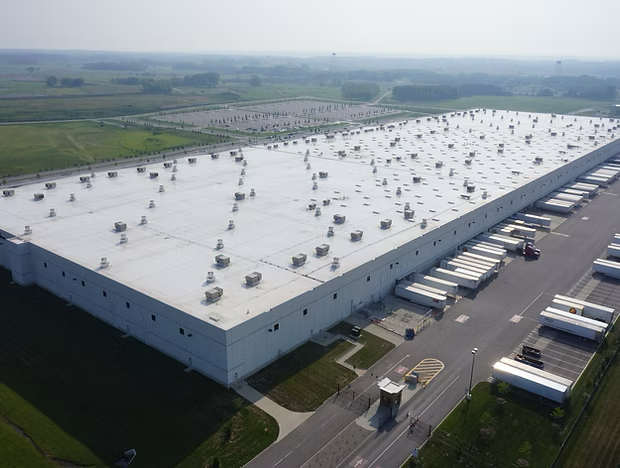 Aerial view of a large white warehouse with rooftop vents beside roads and loading docks in an industrial area