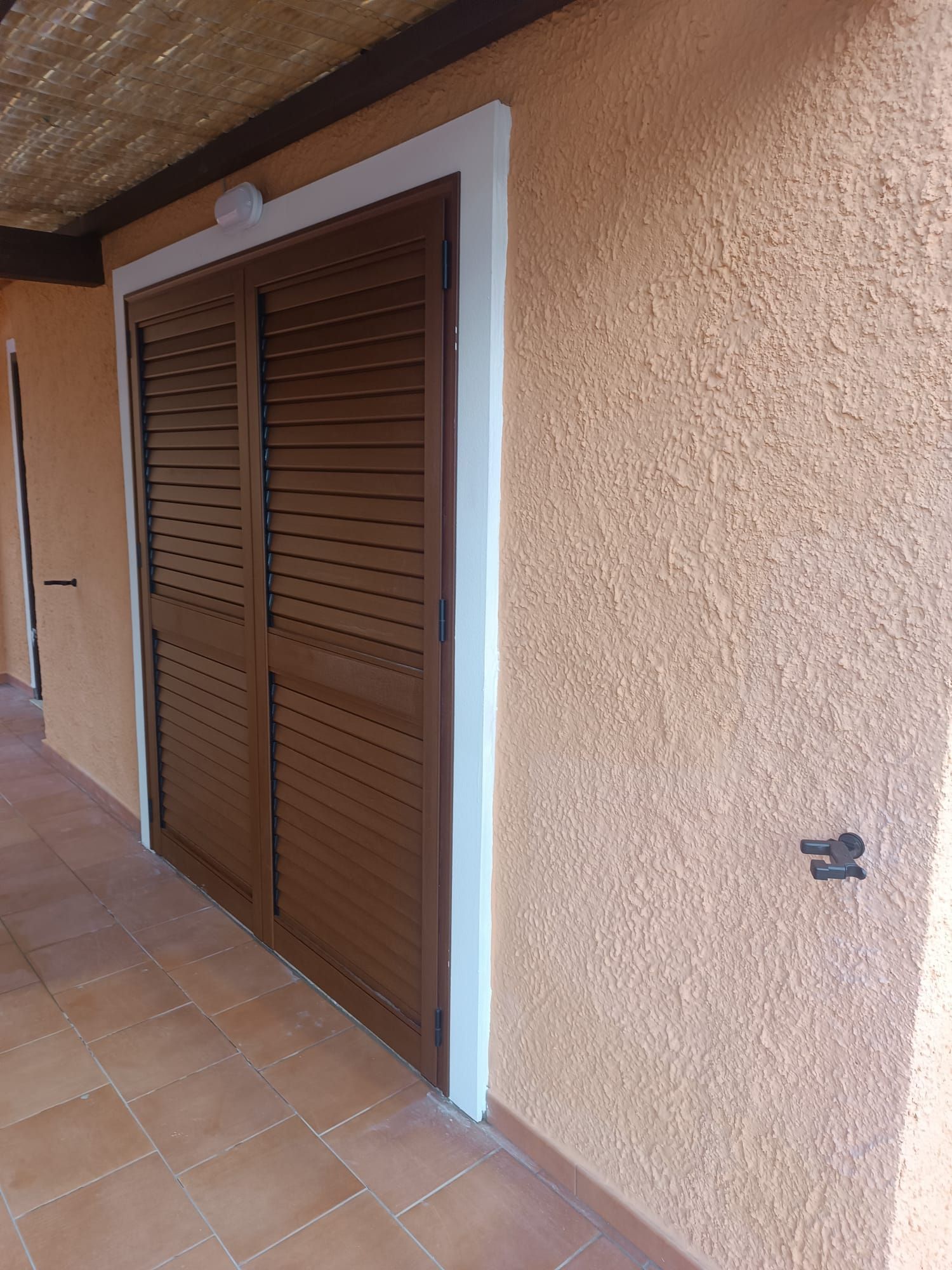 Porte marroni a persiana di un armadio in un corridoio esterno piastrellato, con una parete in stucco