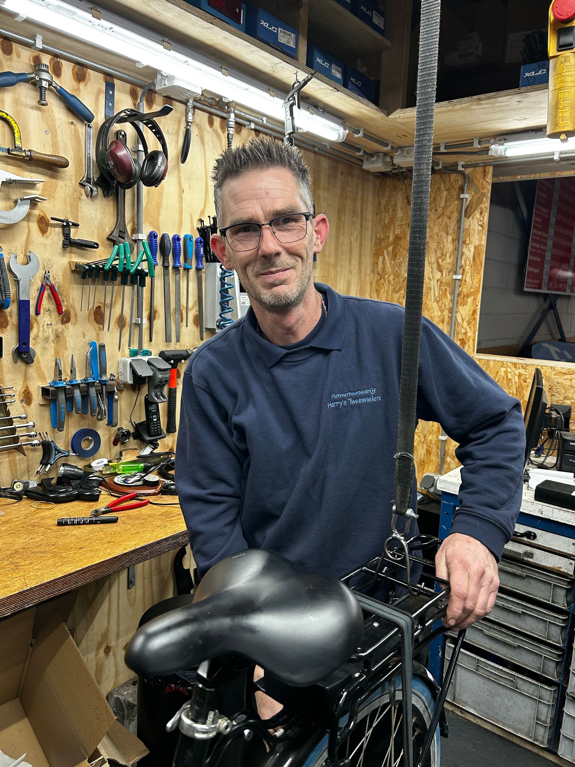 Man in werkplaats die een fietszadel vasthoudt; gereedschap aan de muur.