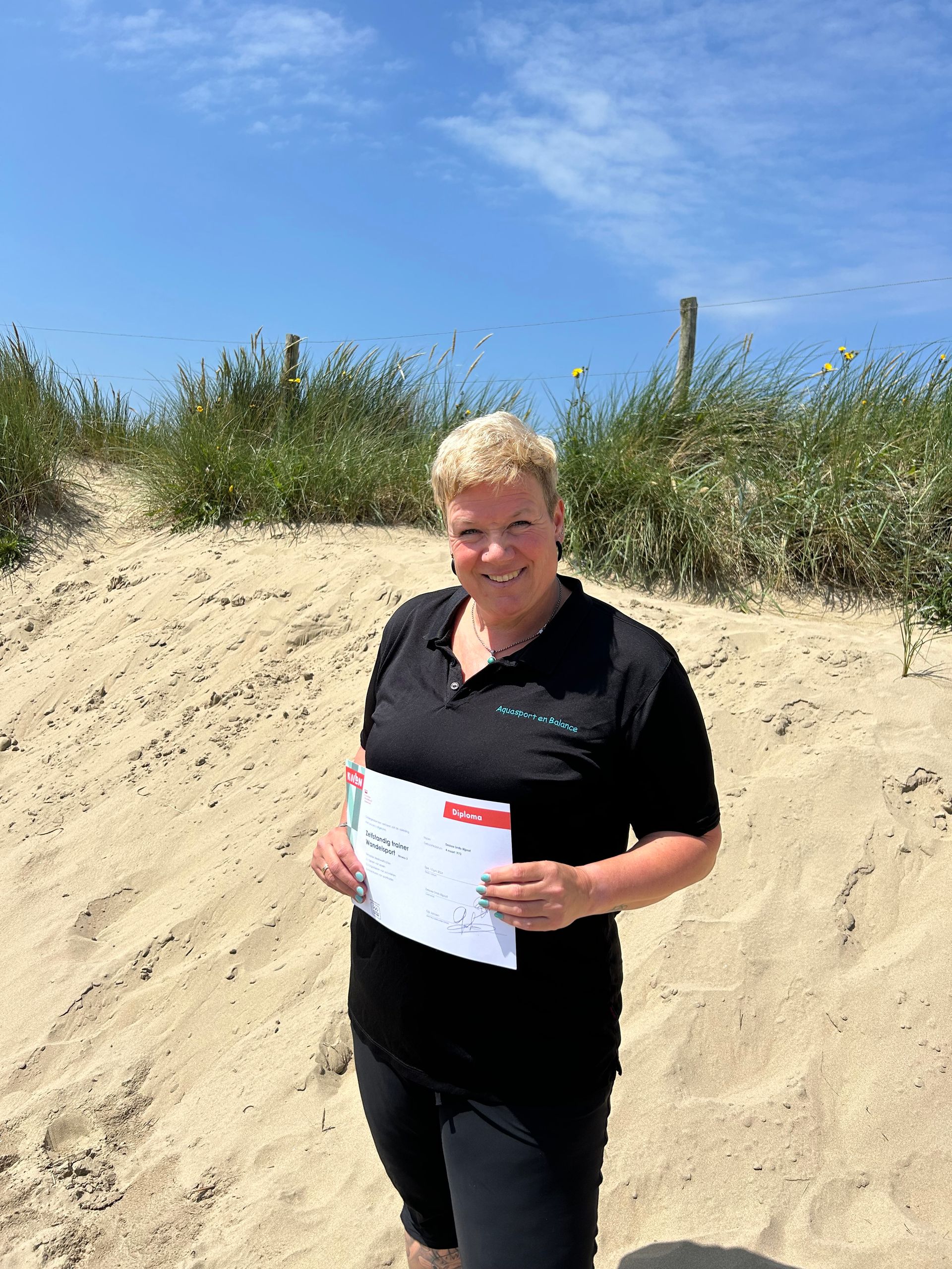 Een vrouw met kort blond haar houdt een vel papier vast en staat op een zandstrand onder een blauwe hemel.