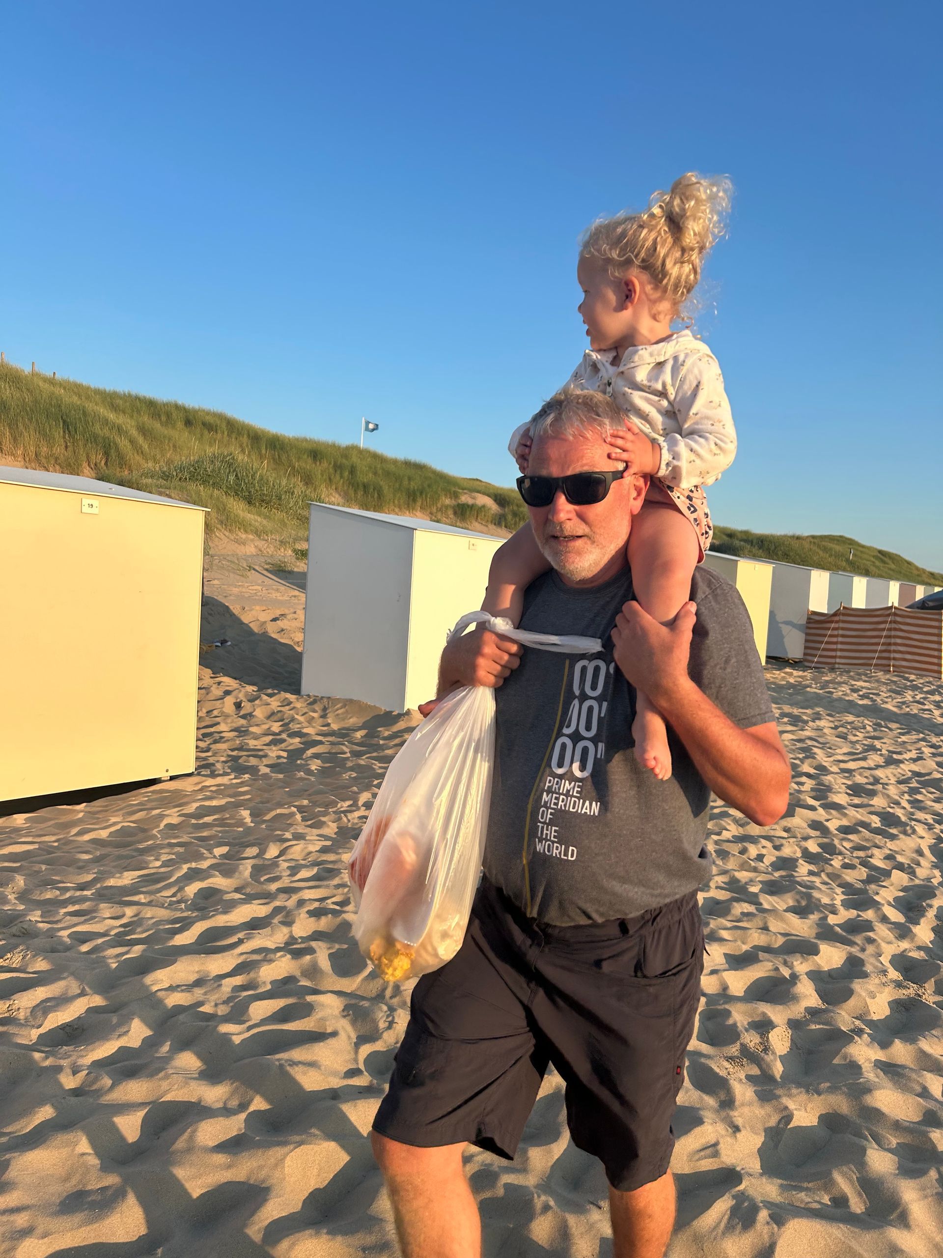 Een man draagt een kind op zijn schouders op het strand, terwijl hij een tas vasthoudt. Ze bevinden zich in de buurt van strandhutten.