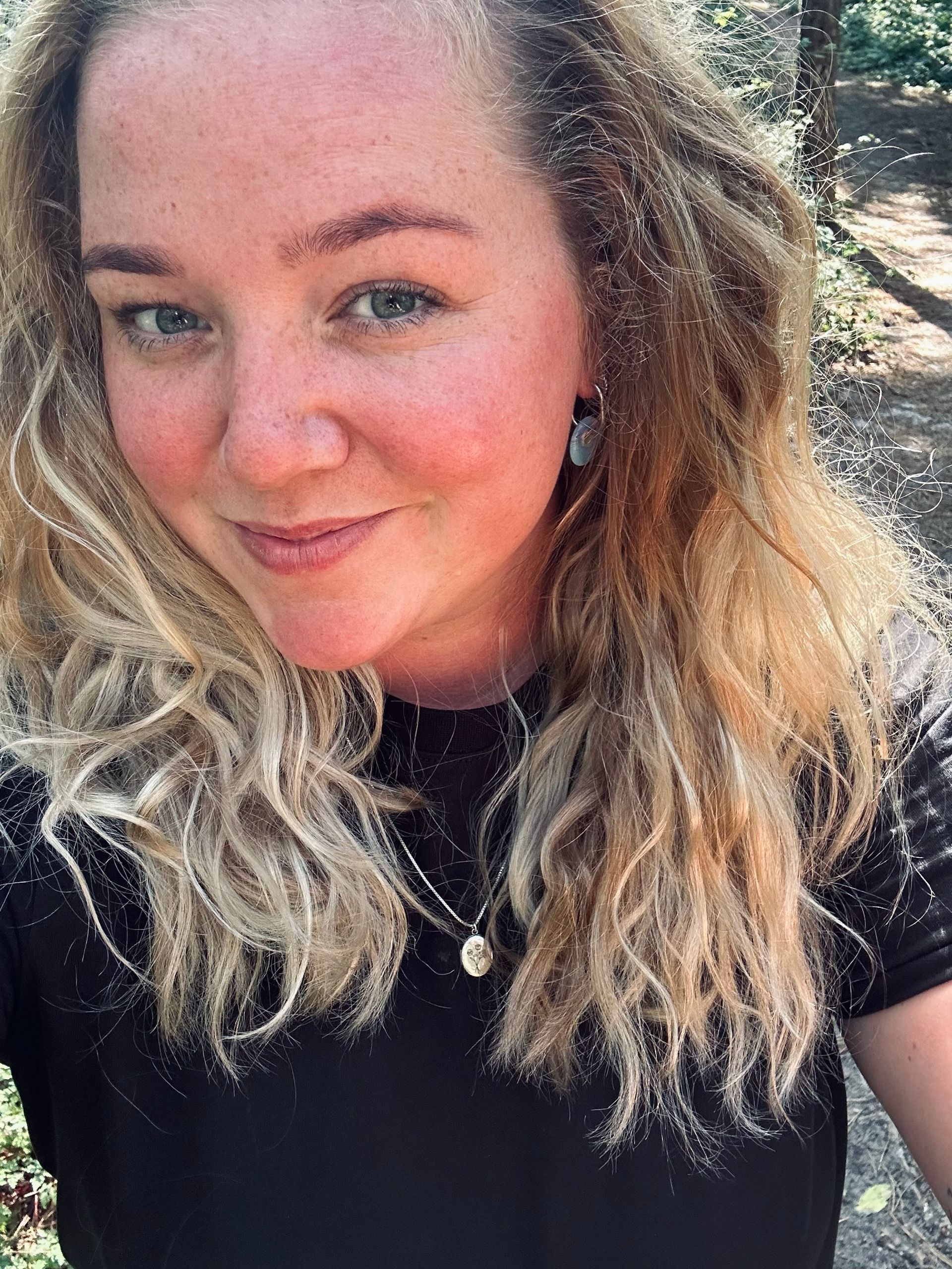 Een vrouw met krullend blond haar lacht naar de camera. Ze draagt een zwart shirt en staat buiten met bomen op de achtergrond.