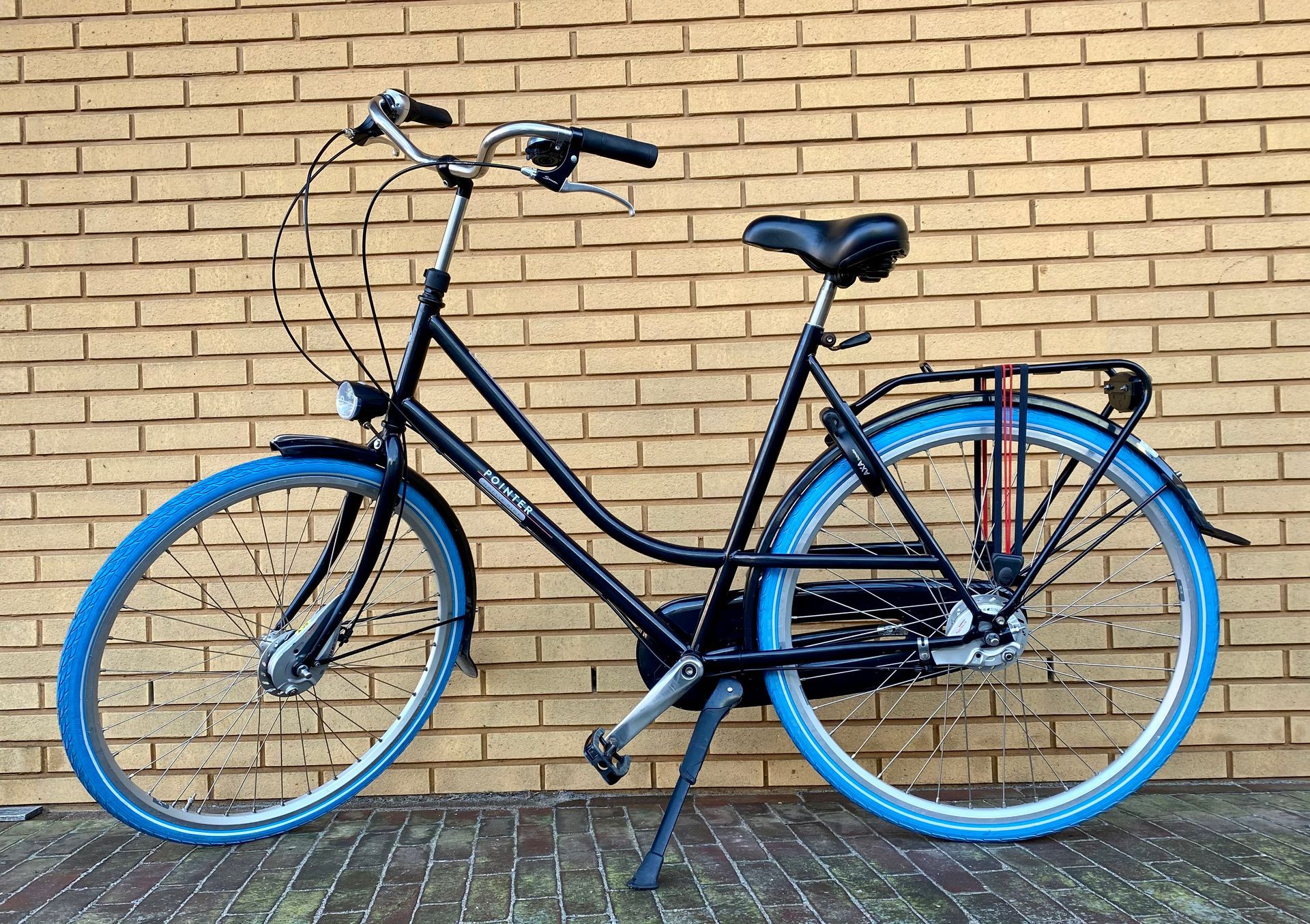 Zwarte fiets met blauwe banden, standaard uitgeklapt, tegen een bakstenen muur.