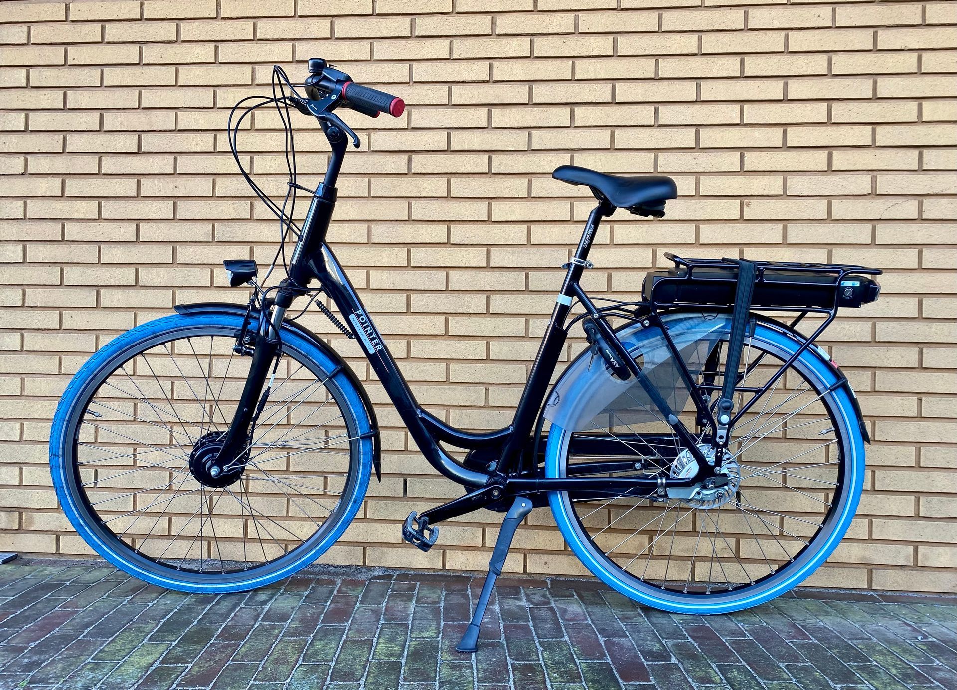 Zwarte elektrische fiets met blauwe banden geparkeerd tegen een bakstenen muur.