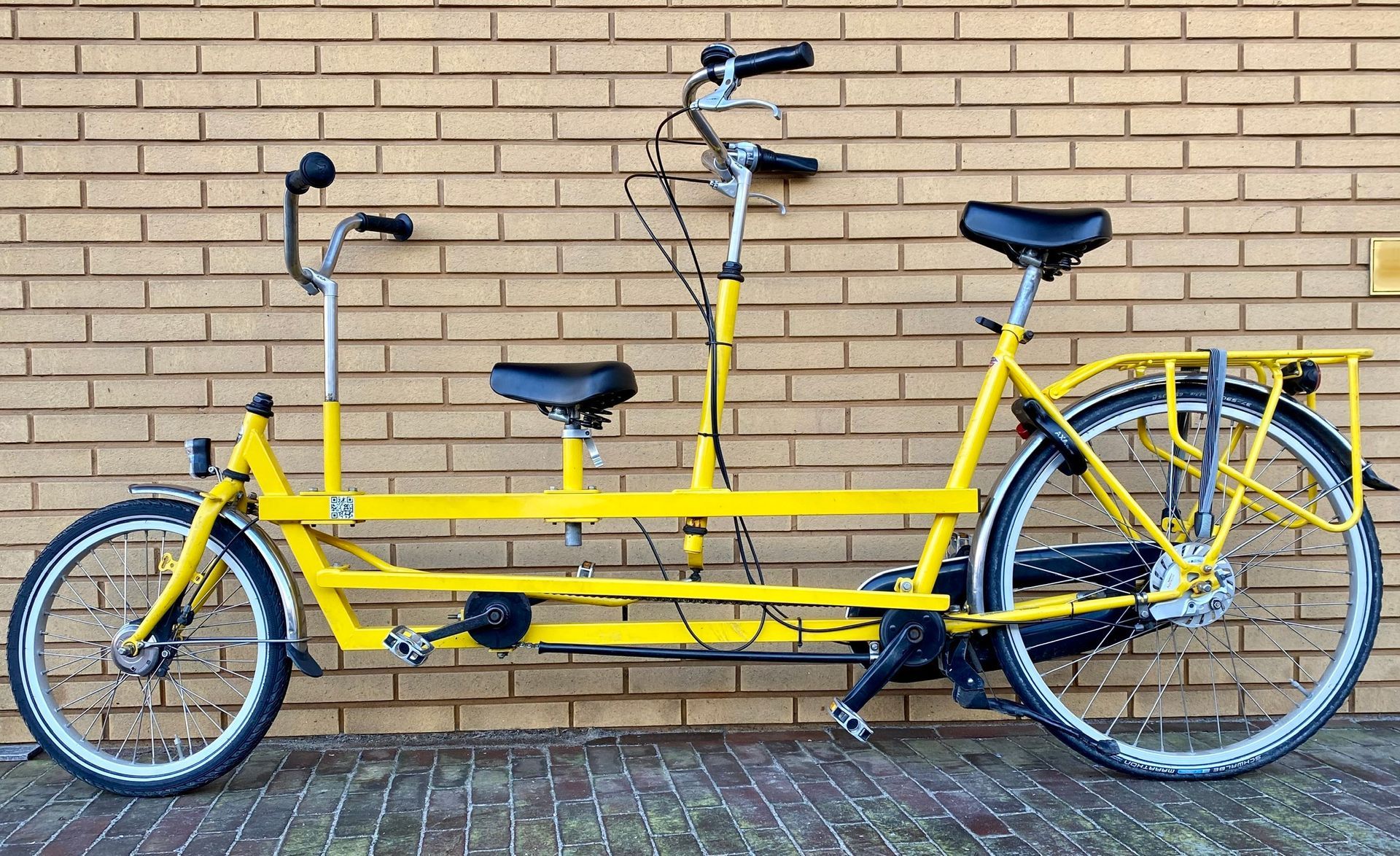 Gele tandemfiets tegen een bakstenen muur. Hij heeft twee zitjes, een stuur en een bagagedrager.