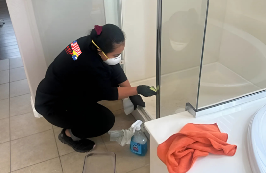 A woman wearing a mask is cleaning a shower door in a bathroom.