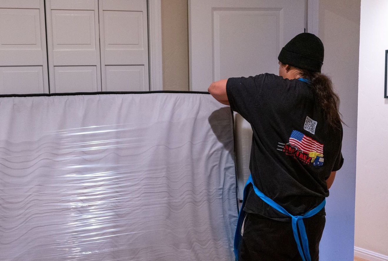 A woman in a black shirt and blue apron is wrapping a mattress in plastic wrap.