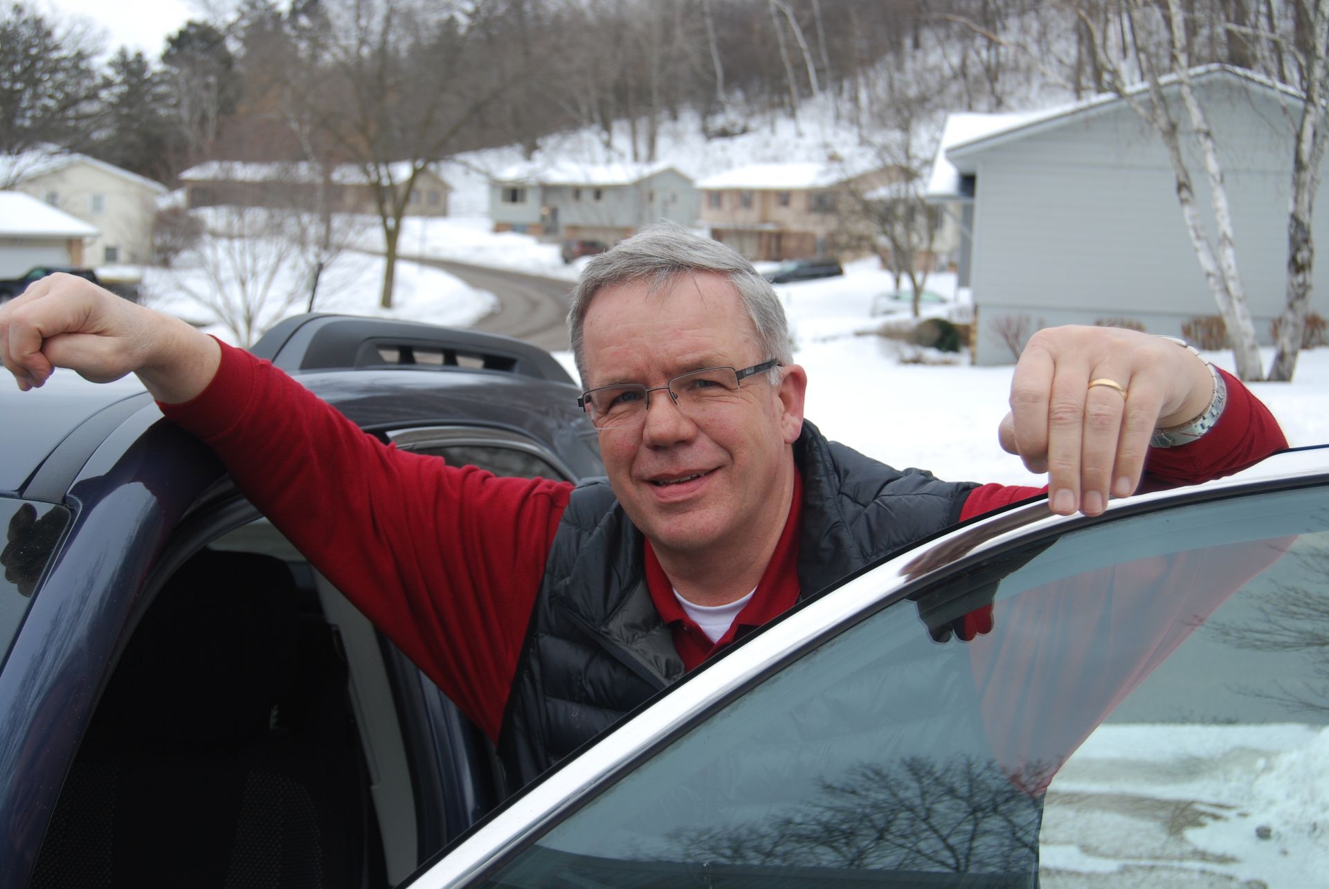 Man leans against a car in a snowy residential area. He wears glasses, a red shirt, and a vest, smiling.