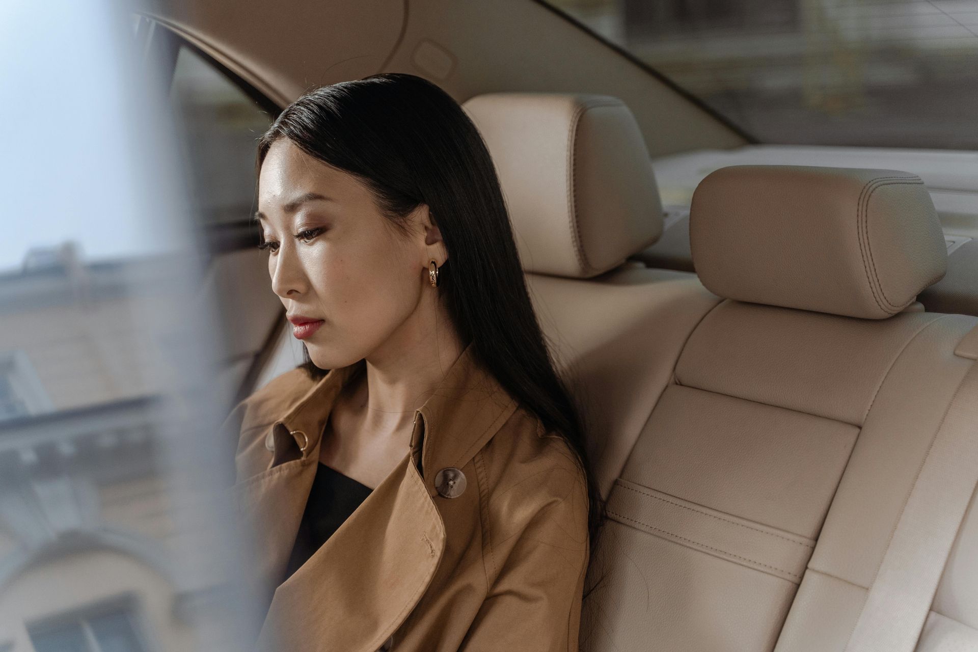 Woman in tan coat sits in a car, looking down. Beige interior, window visible.