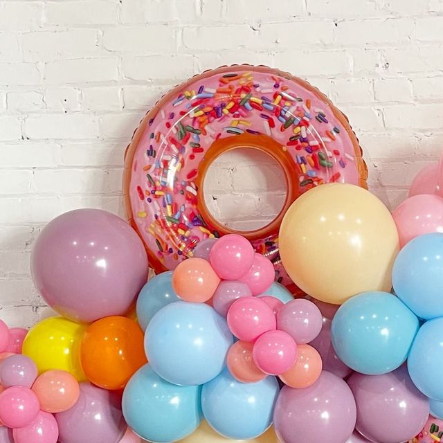 A pink donut with sprinkles is surrounded by balloons.