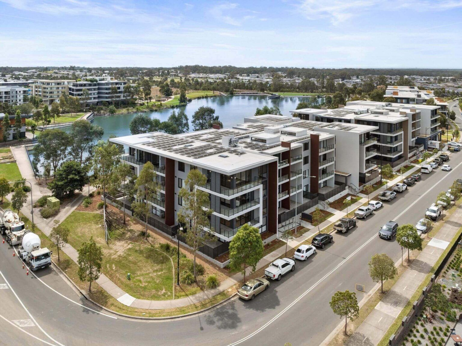 Aerial view of modern apartment buildings next to a lake and road, with parked cars and trees.