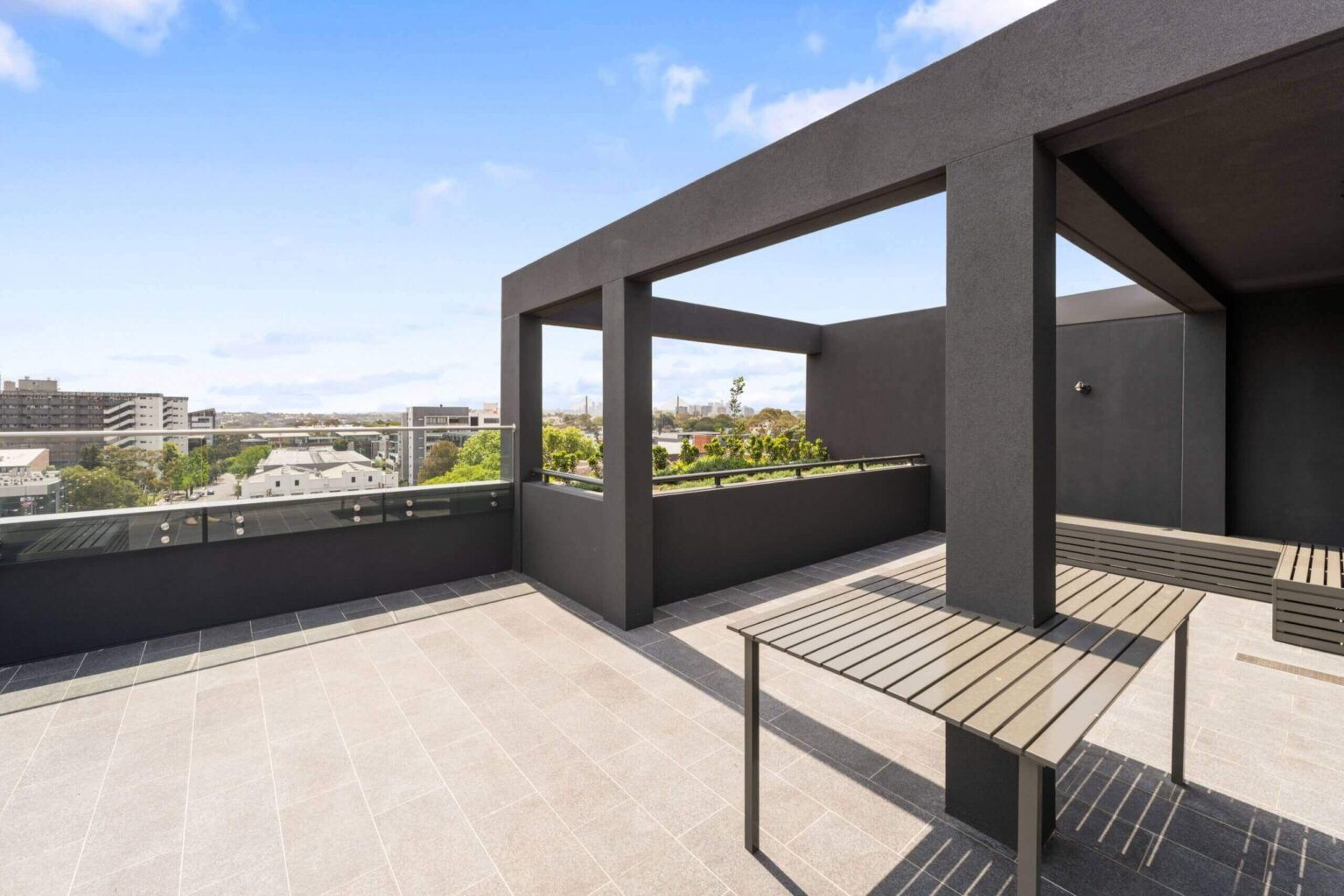 Rooftop patio with dark gray walls, light gray floor, and wooden tables, overlooking a city.