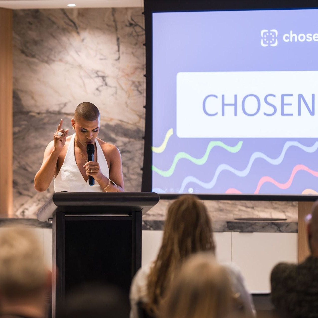 Person speaking at a podium, gesturing, in front of a screen with the word