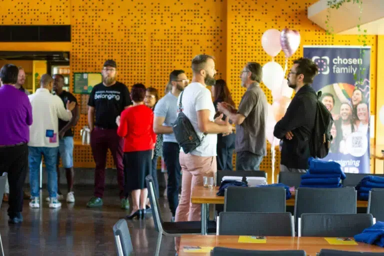 People gathered in a room, conversing near tables and a banner. Yellow textured wall in background, some balloons.