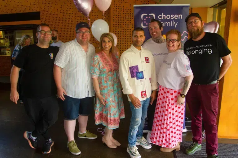 Group of seven people smiling in front of a “Chosen Family” banner, with balloons.