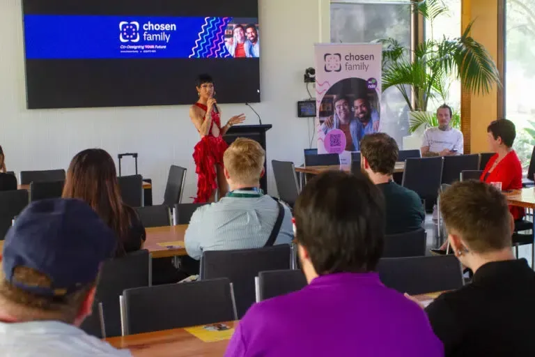 Woman speaking at a Chosen Family event, in front of an audience, a screen, and a banner.