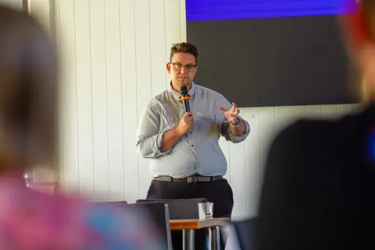 Man speaking into a microphone, gesturing with his hands. He is wearing glasses and a button-down shirt.