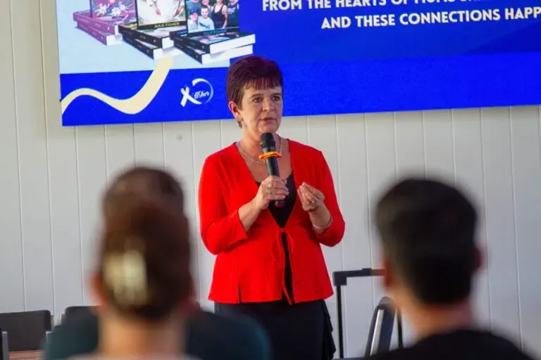 Woman in red jacket speaking into a microphone, addressing a seated audience.