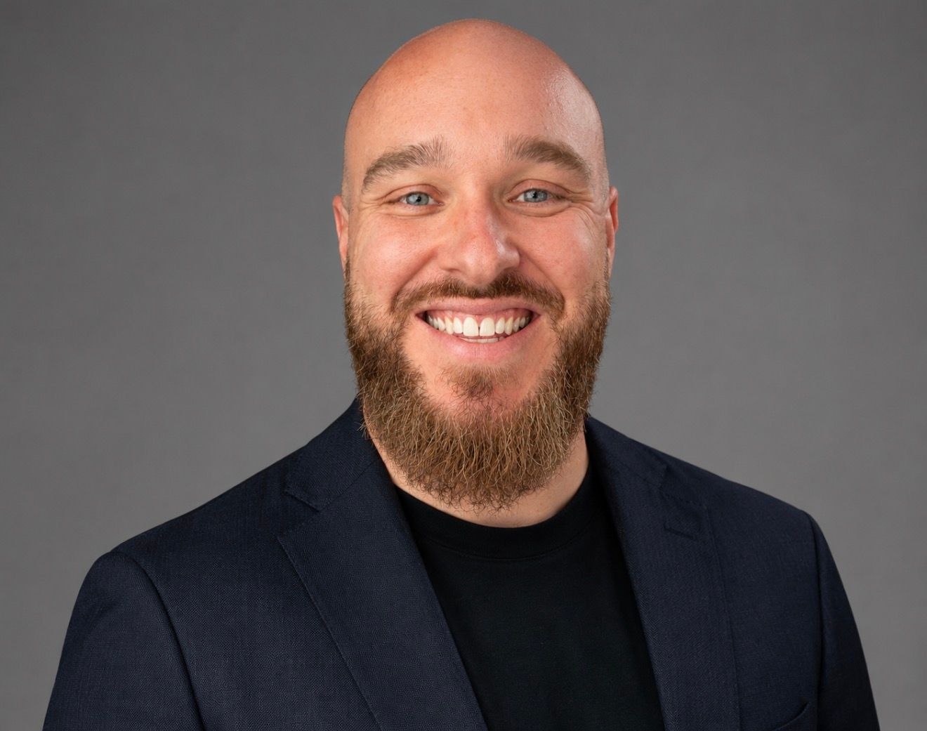 Smiling bald man with a beard in a dark blazer and black shirt against a gray background