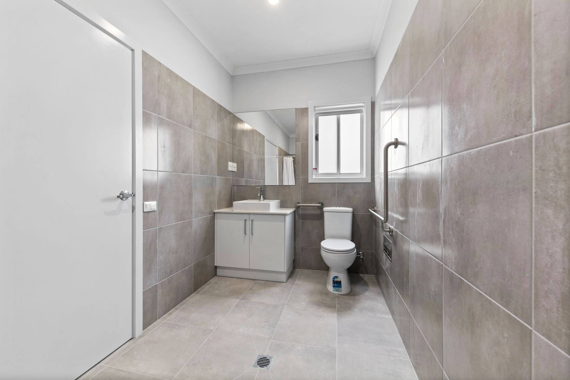 Accessible bathroom with grey tiled walls and floor, toilet with grab bar, vanity, and window.