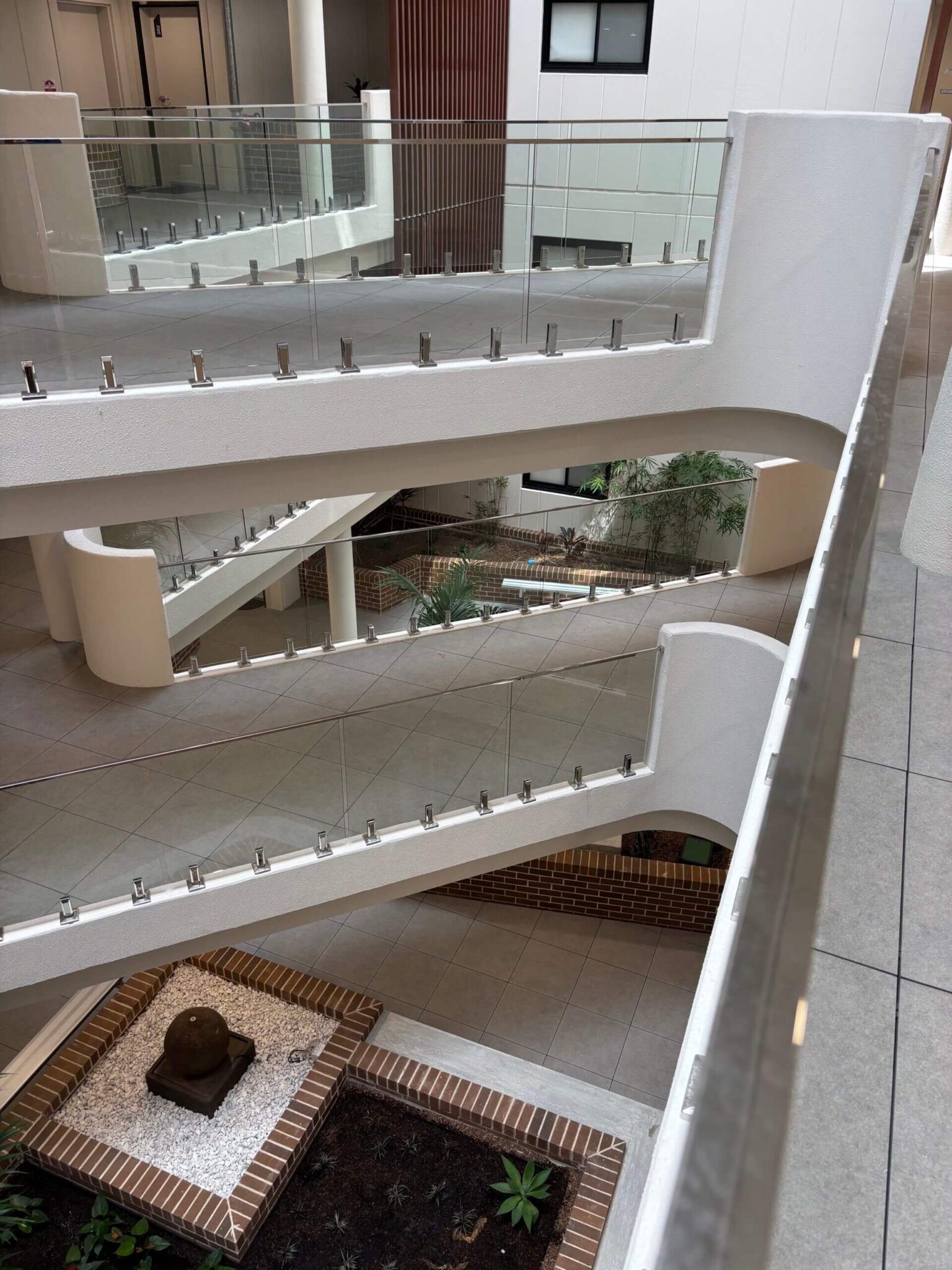 Multi-level indoor building with walkways and glass railings overlooking a garden area with plants and decorative stones.