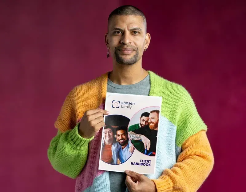 Man holding a brochure with photos of smiling people; wearing a colorful sweater in front of a magenta background.