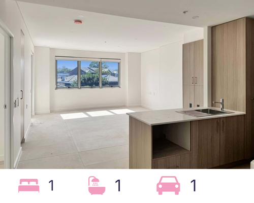 The remaining modern apartment interior at Leumeah Apartments features white walls and tiled floors. An open-plan kitchen with wooden cabinets stands to the right, while large windows ahead allow natural light to fill this perfect space for single occupancy.