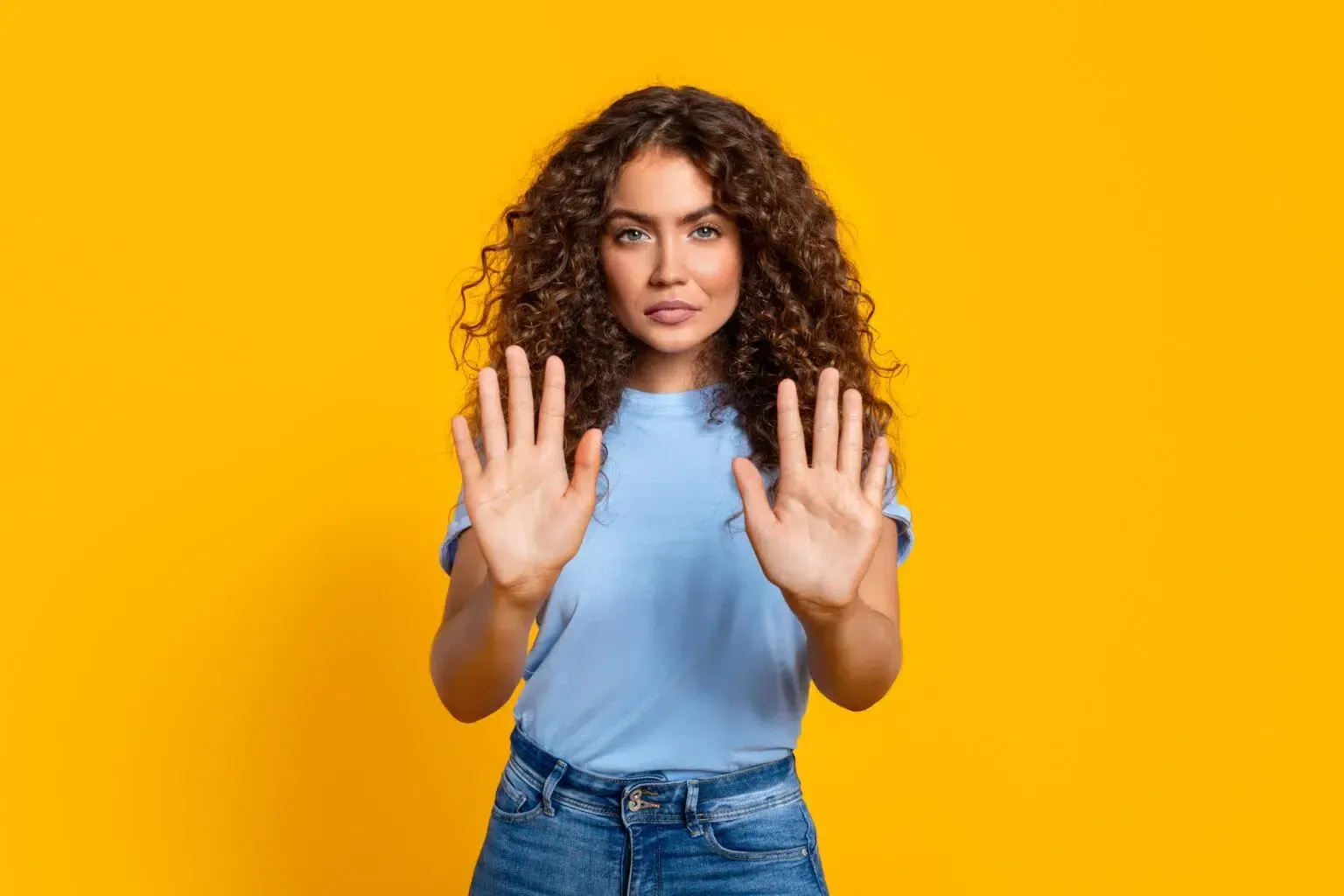 Woman with curly brown hair, blue shirt, and jeans, hands up to set boundaries and is against a yellow background.