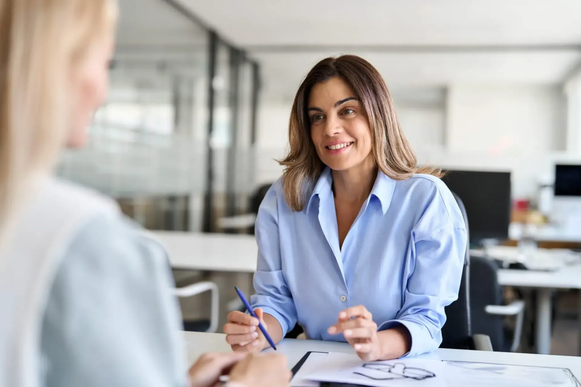 Woman in blue shirt smiles, talking to another person at a desk in an office setting.