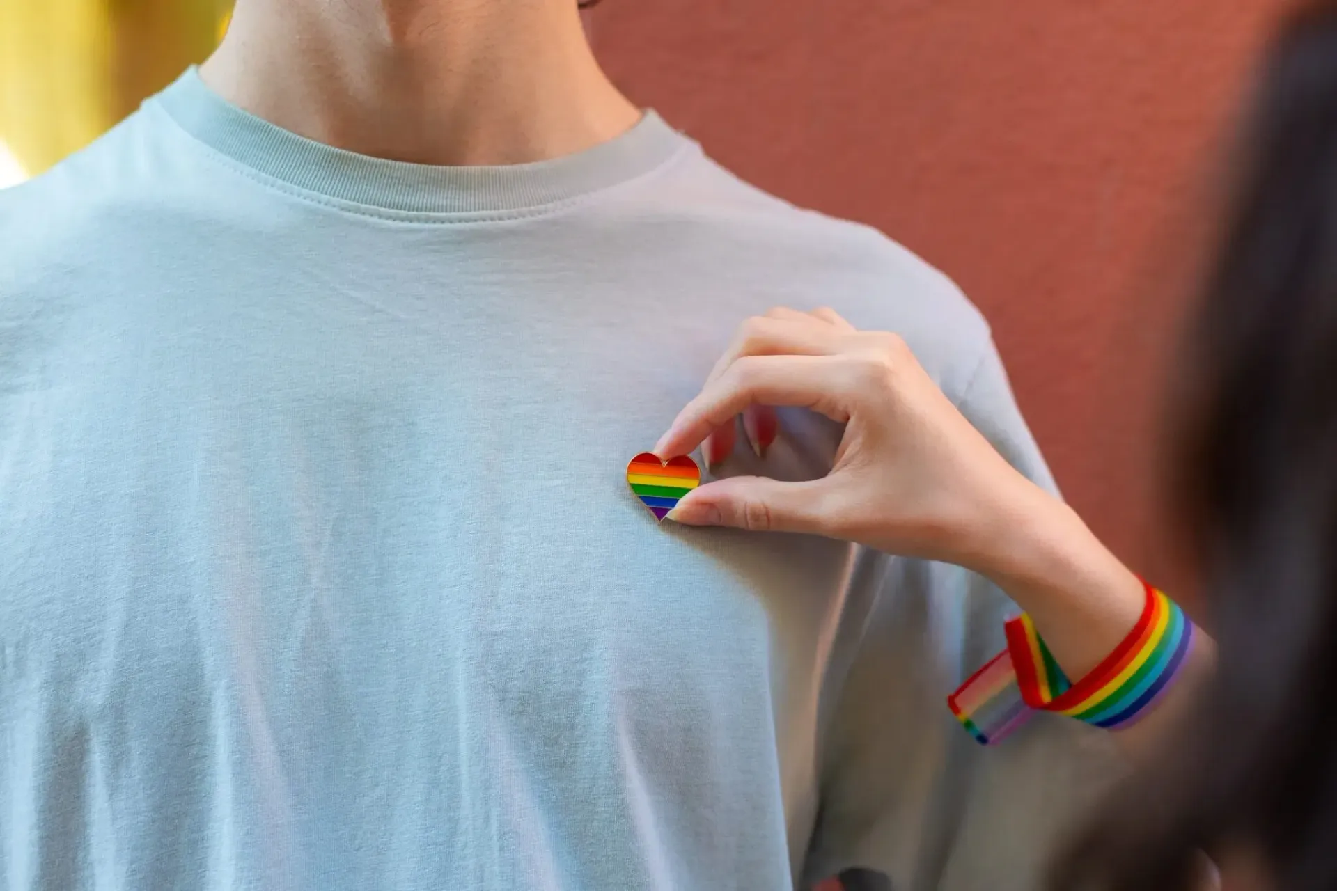 Hand pinning a rainbow heart badge on a person's shirt; rainbow bracelet visible.