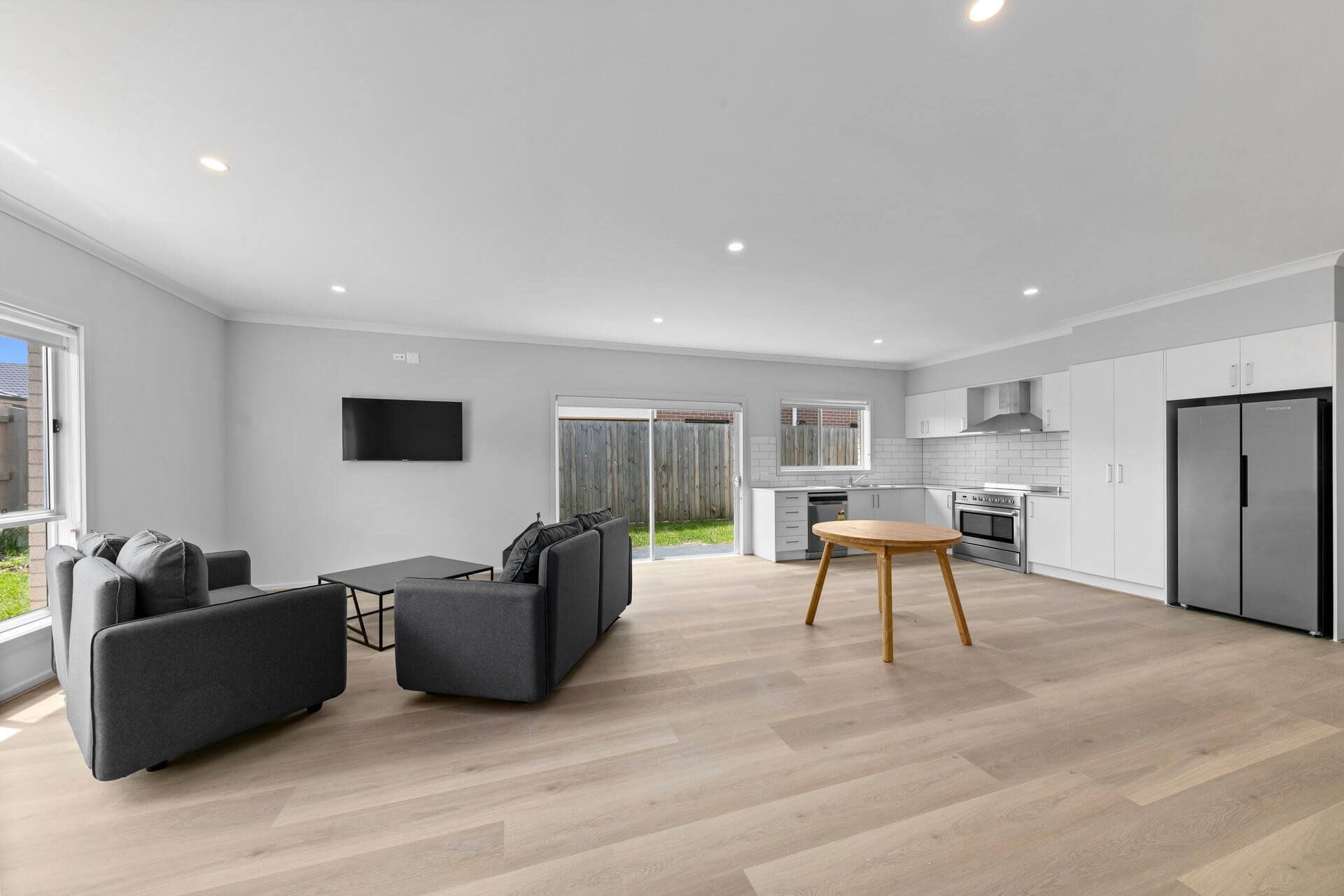 Open-plan living space with grey sofas, kitchen, light wood flooring, and access to a backyard.