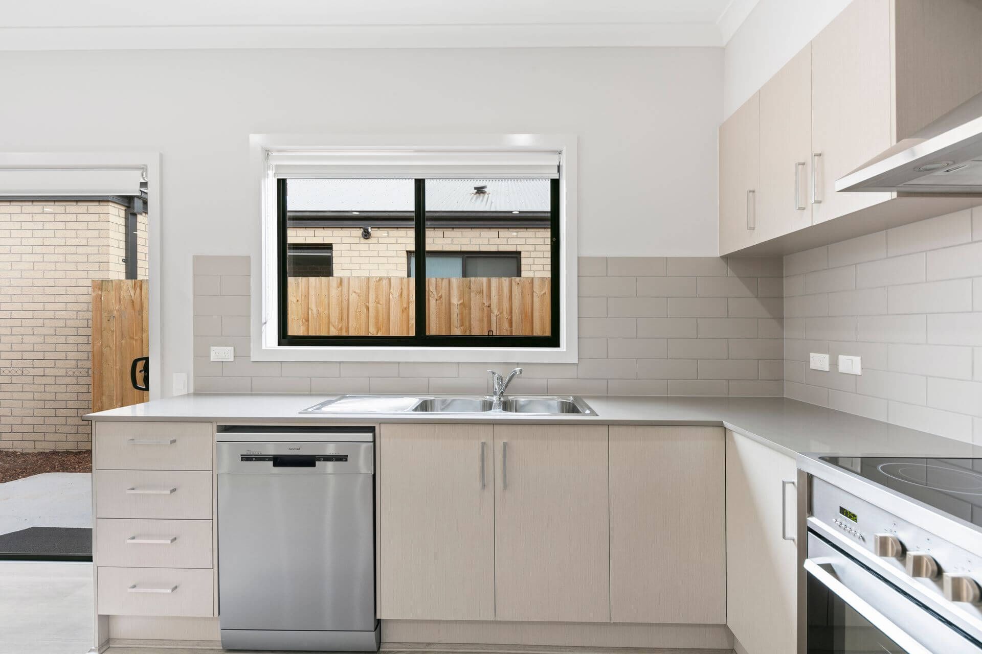 Kitchen with light-colored cabinets, stainless steel dishwasher and sink, and a window overlooking a fence.