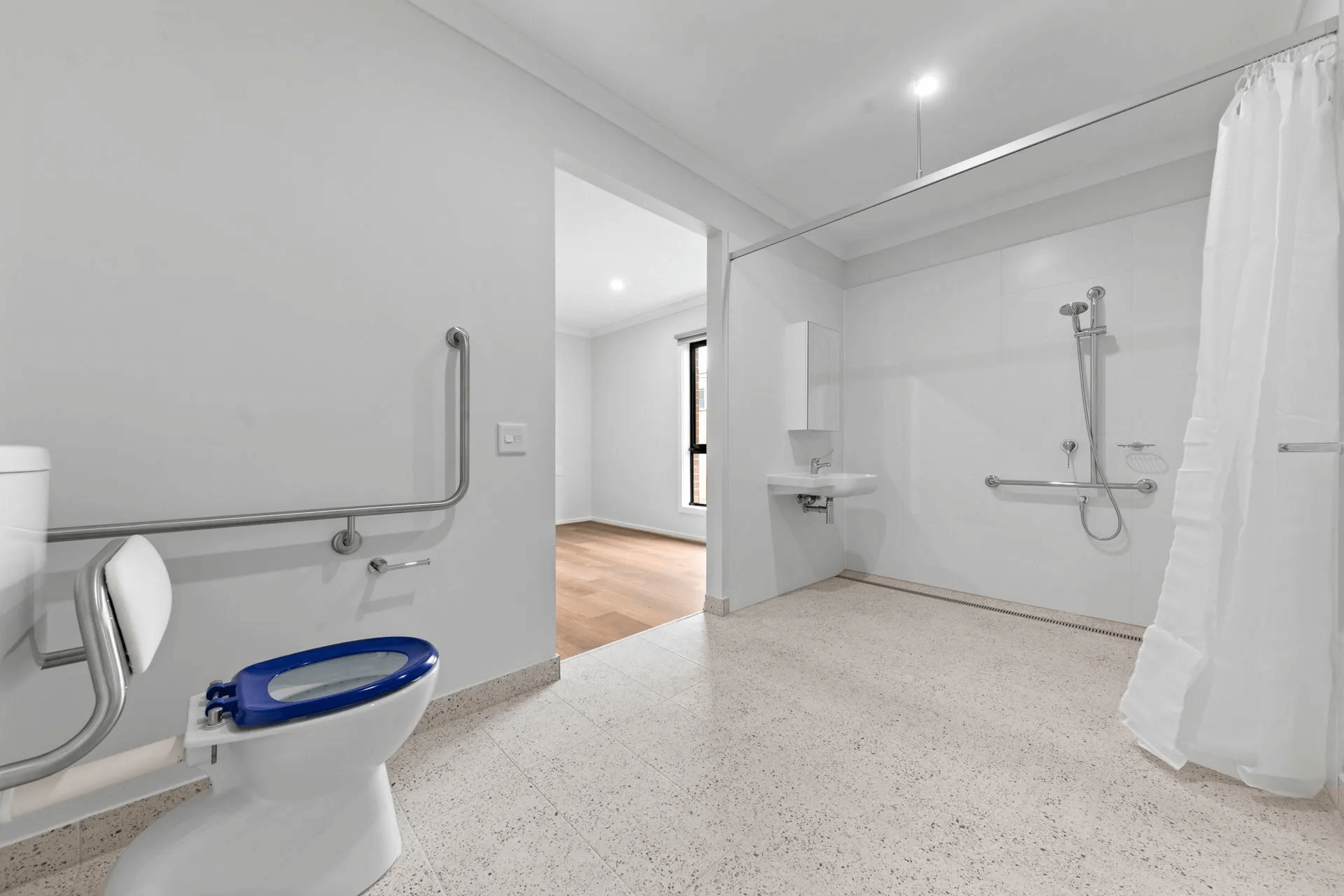 Accessible bathroom with roll-in shower, grab bars, and accessible toilet. White walls and speckled flooring.