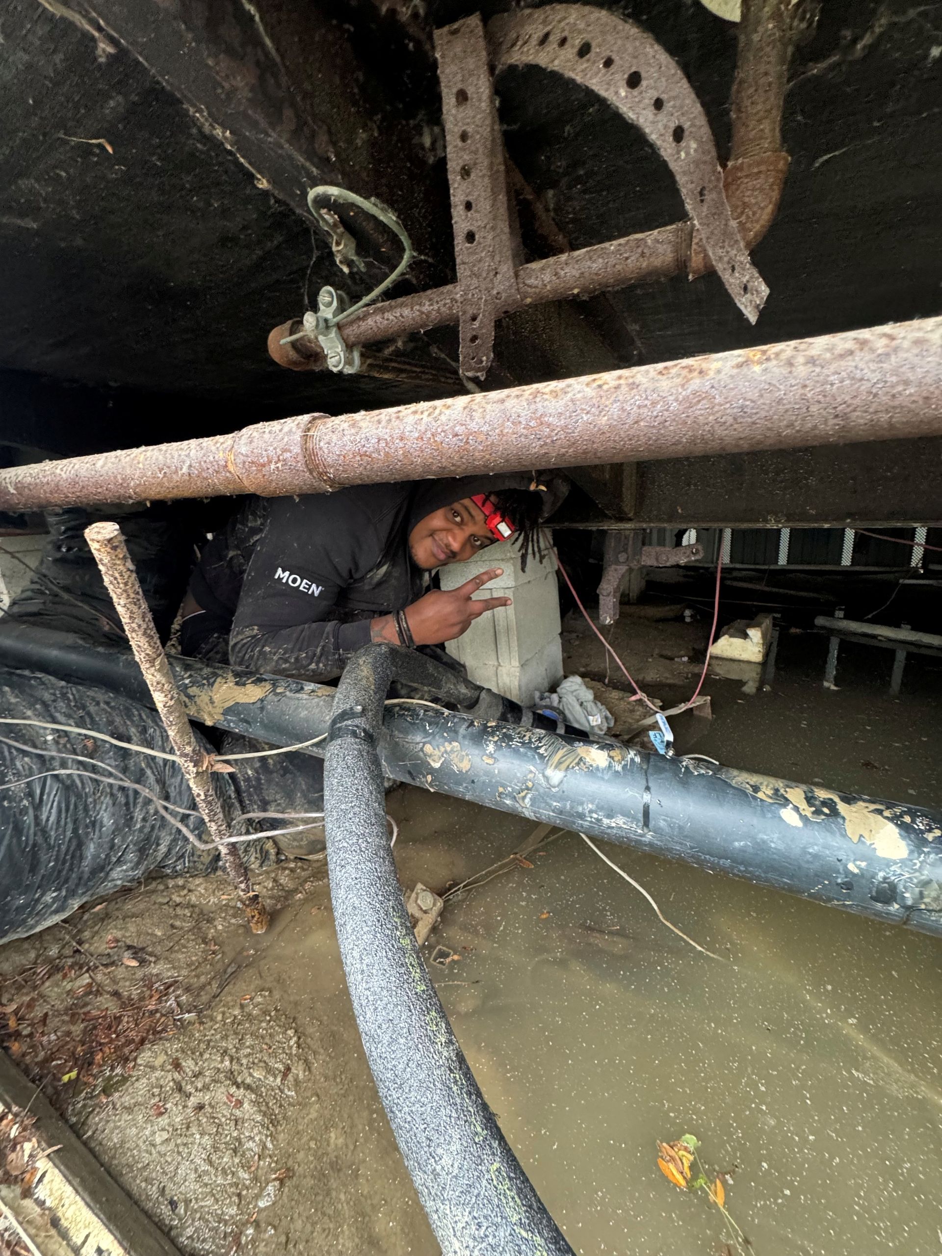 Worker crouches under industrial machinery, handling a hose in a tight, dirty maintenance area.