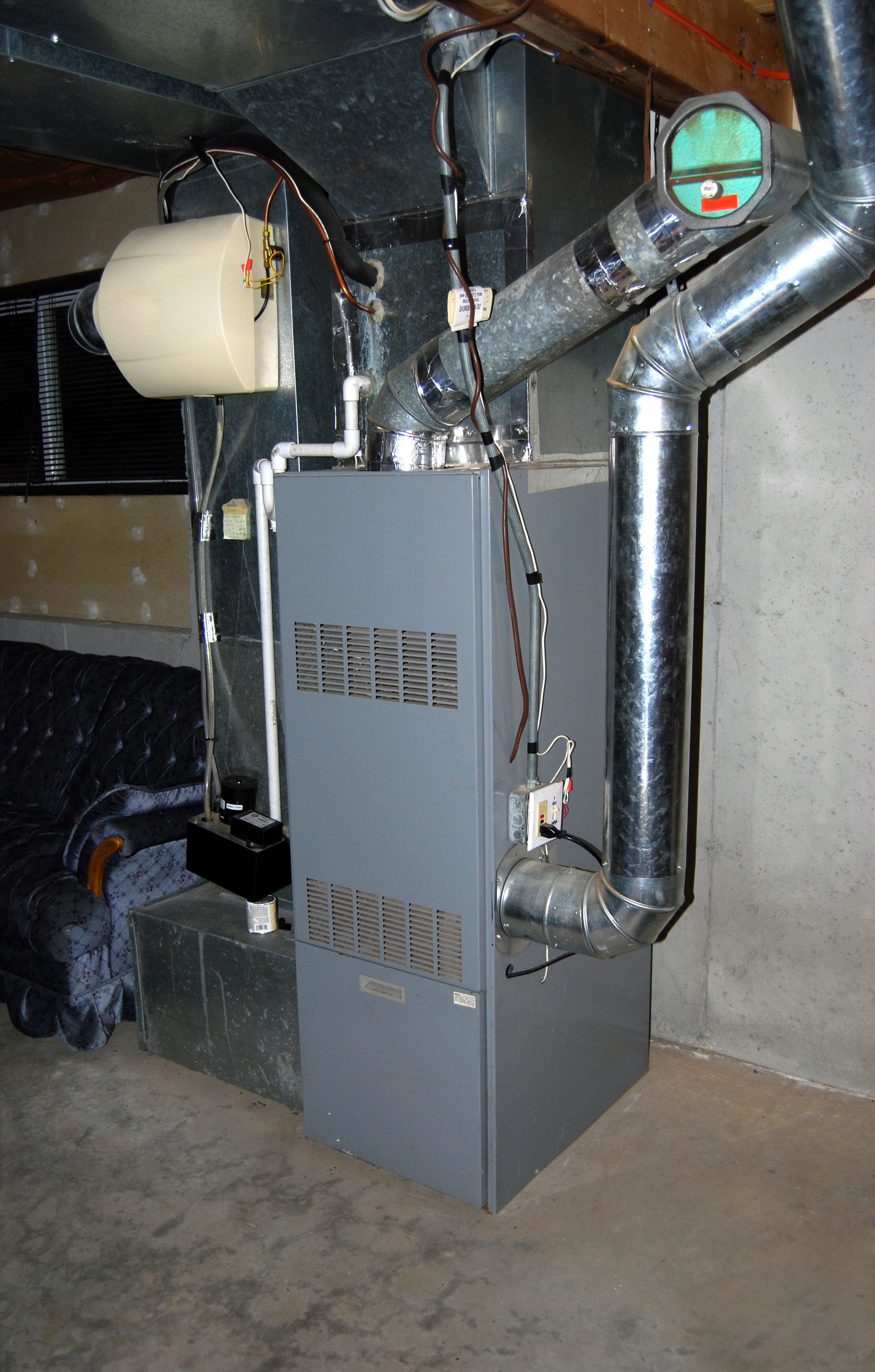 Basement furnace with silver ductwork and a white expansion tank against a concrete wall.