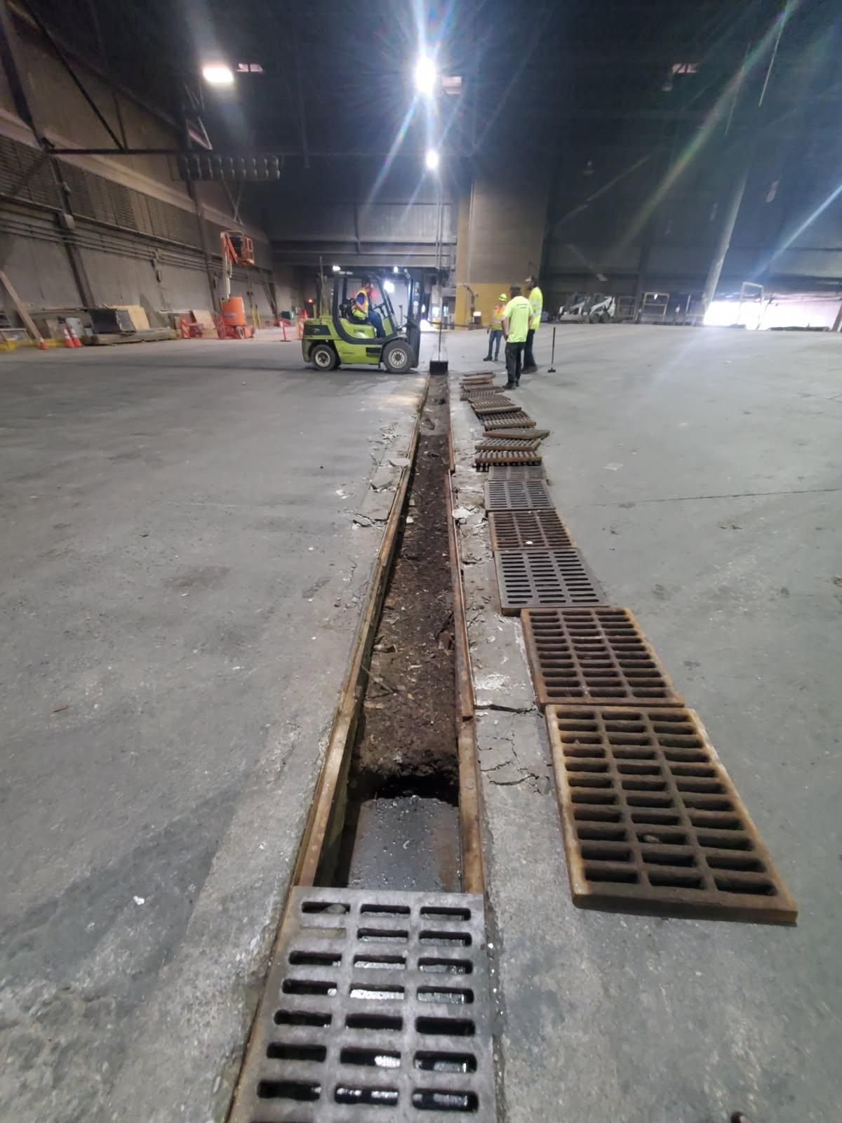 Industrial warehouse floor with an open drainage trench and workers near a forklift in the distance