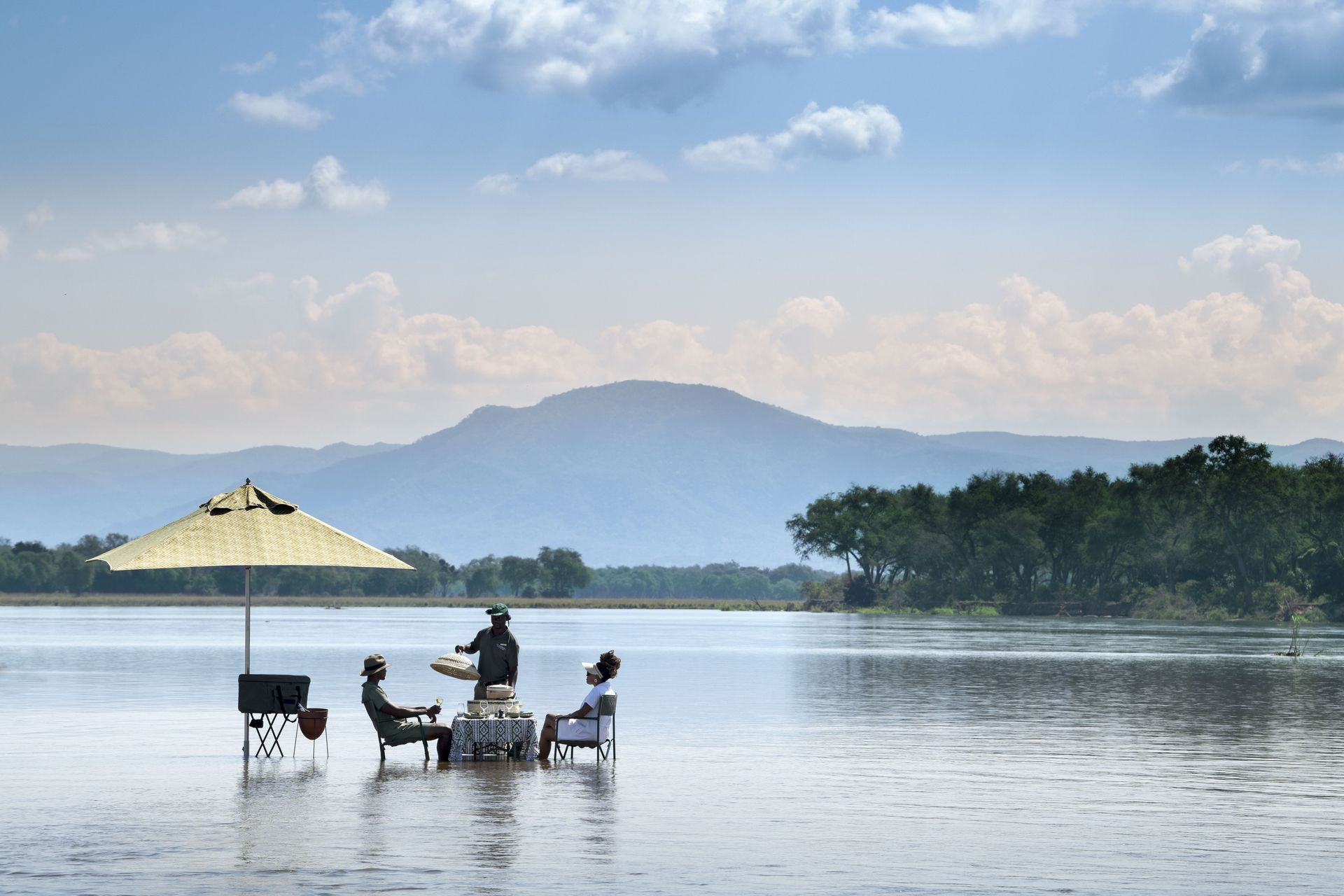 Three people sit at a table under a parasol, partially submerged in a calm body of water with a mountain range in distance.
