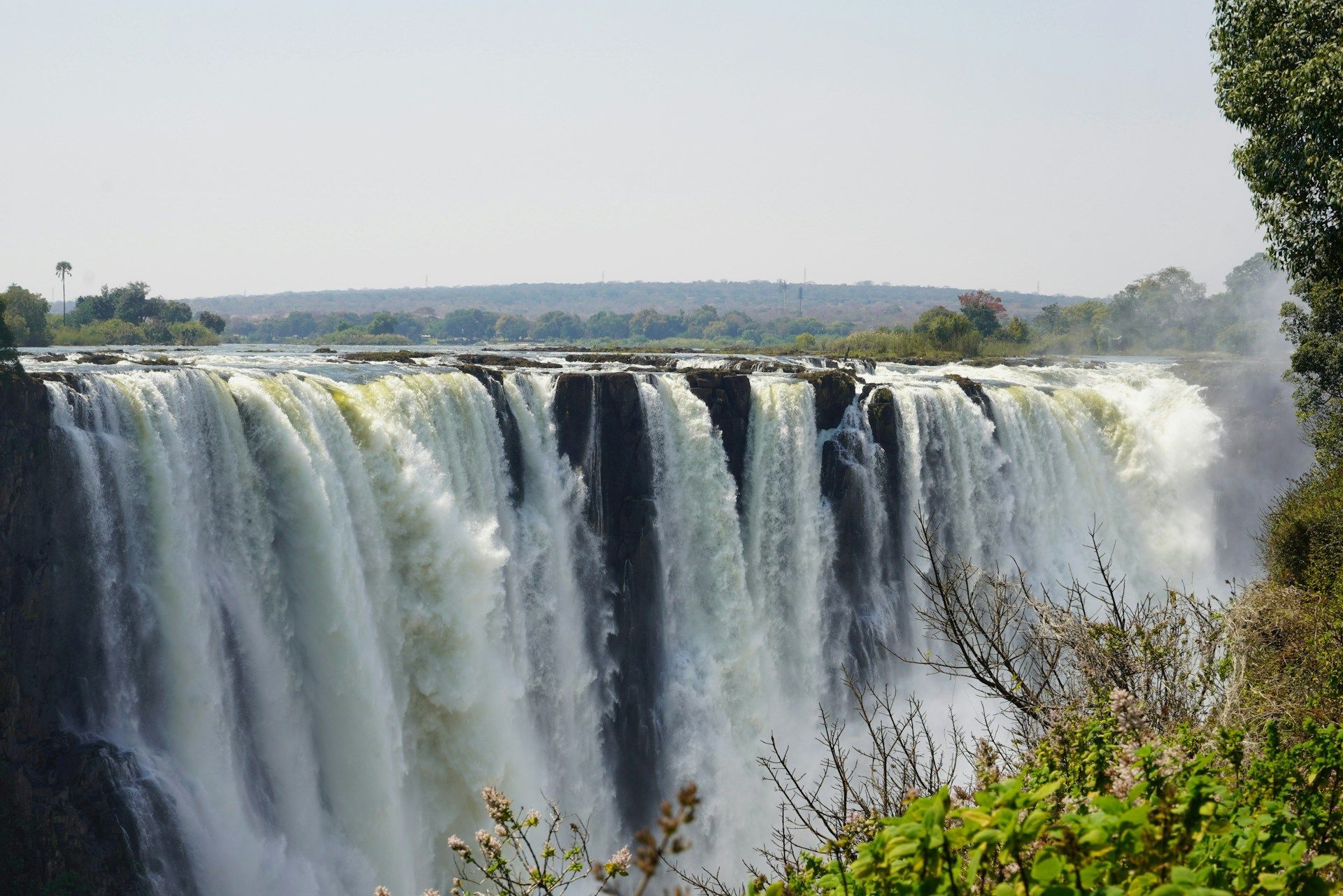 Victoria Falls, a wide, powerful waterfall cascading over a cliff edge amidst lush green vegetation under a clear sky.
