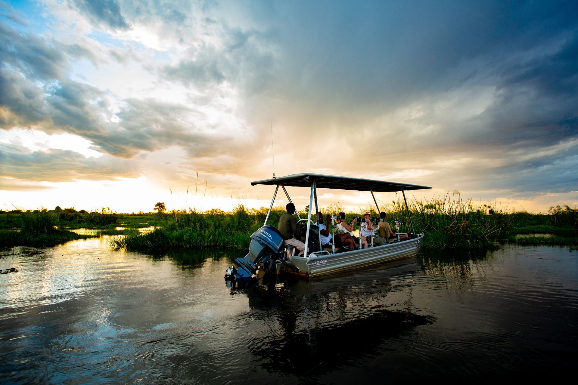 A tour boat filled with passengers travels through calm, marshy waters under a dramatic, cloudy sunset sky.