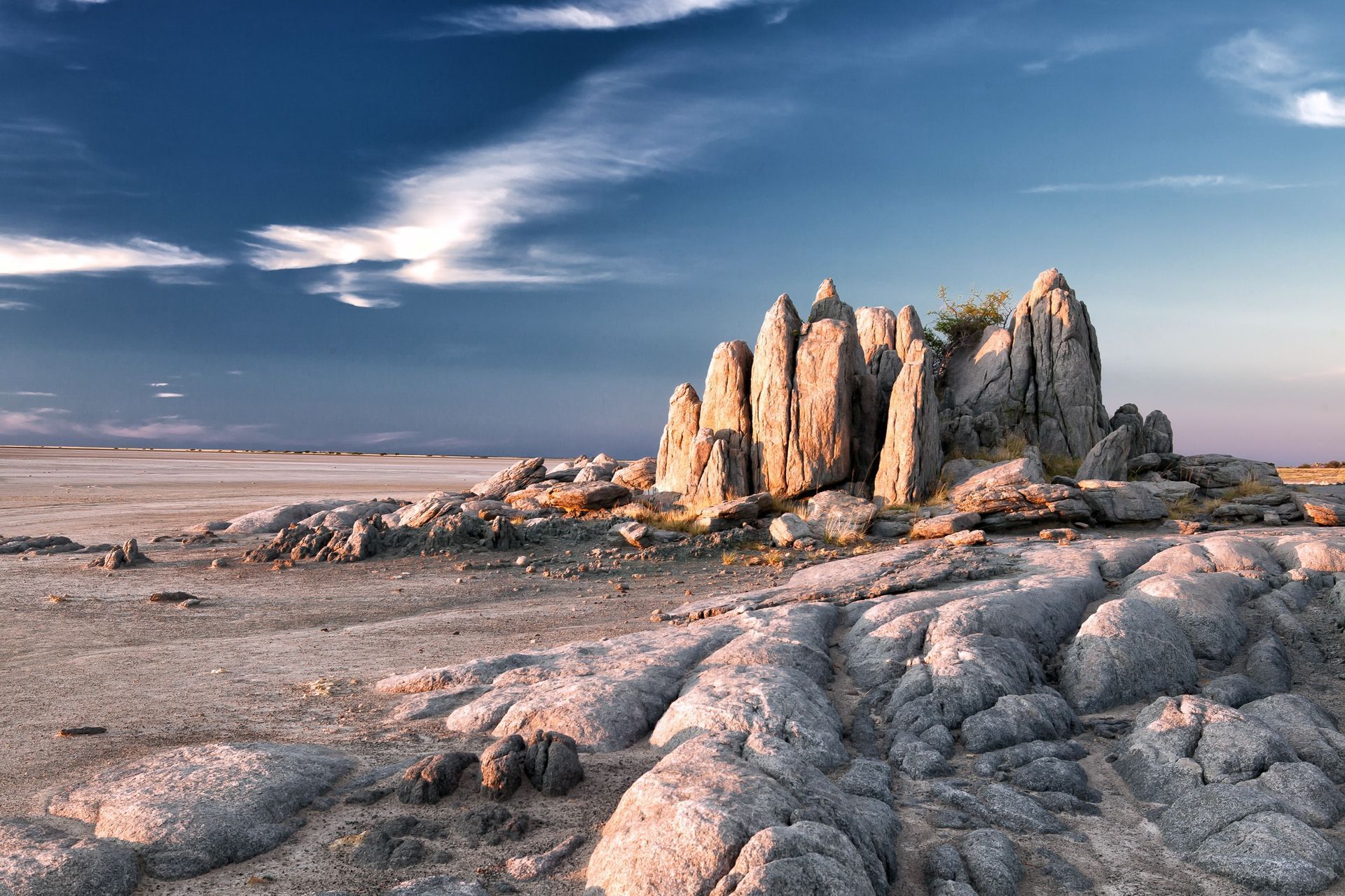 A cluster of craggy, light-colored rock spires stands in a vast, arid, desert-like landscape under a deep blue sky.