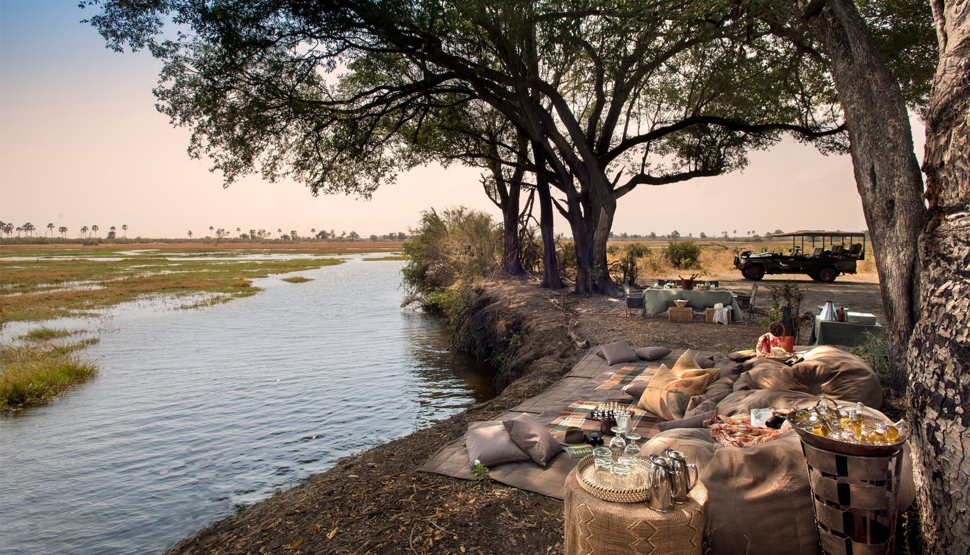 A picnic setup under a large tree overlooking a narrow river in a savanna landscape with a safari vehicle parked nearby.