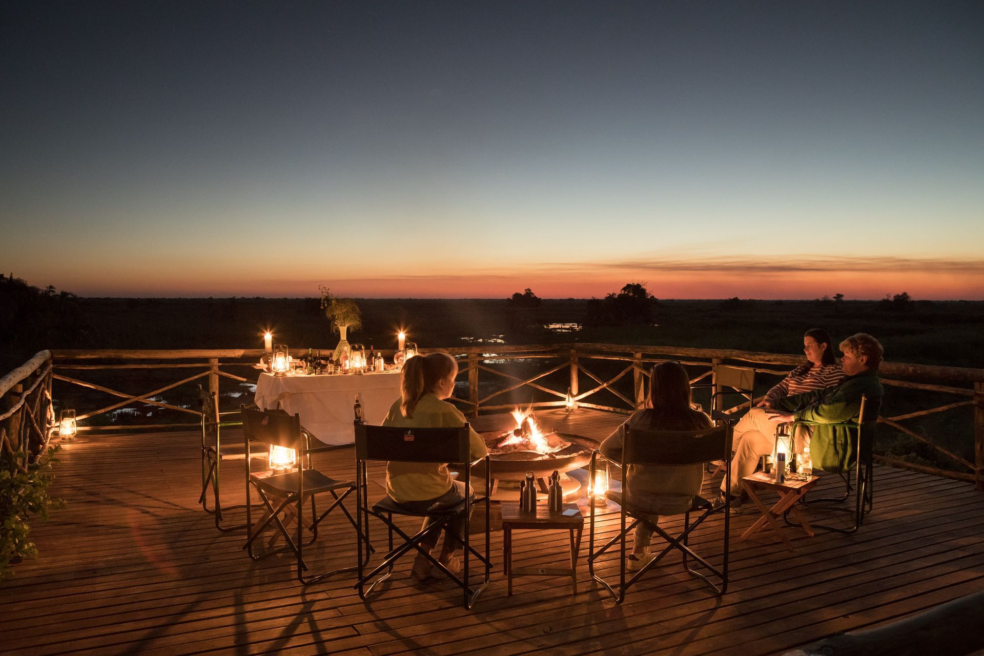 Four people relax on a wooden deck around a small fire pit at dusk, with a dining table set in the background.