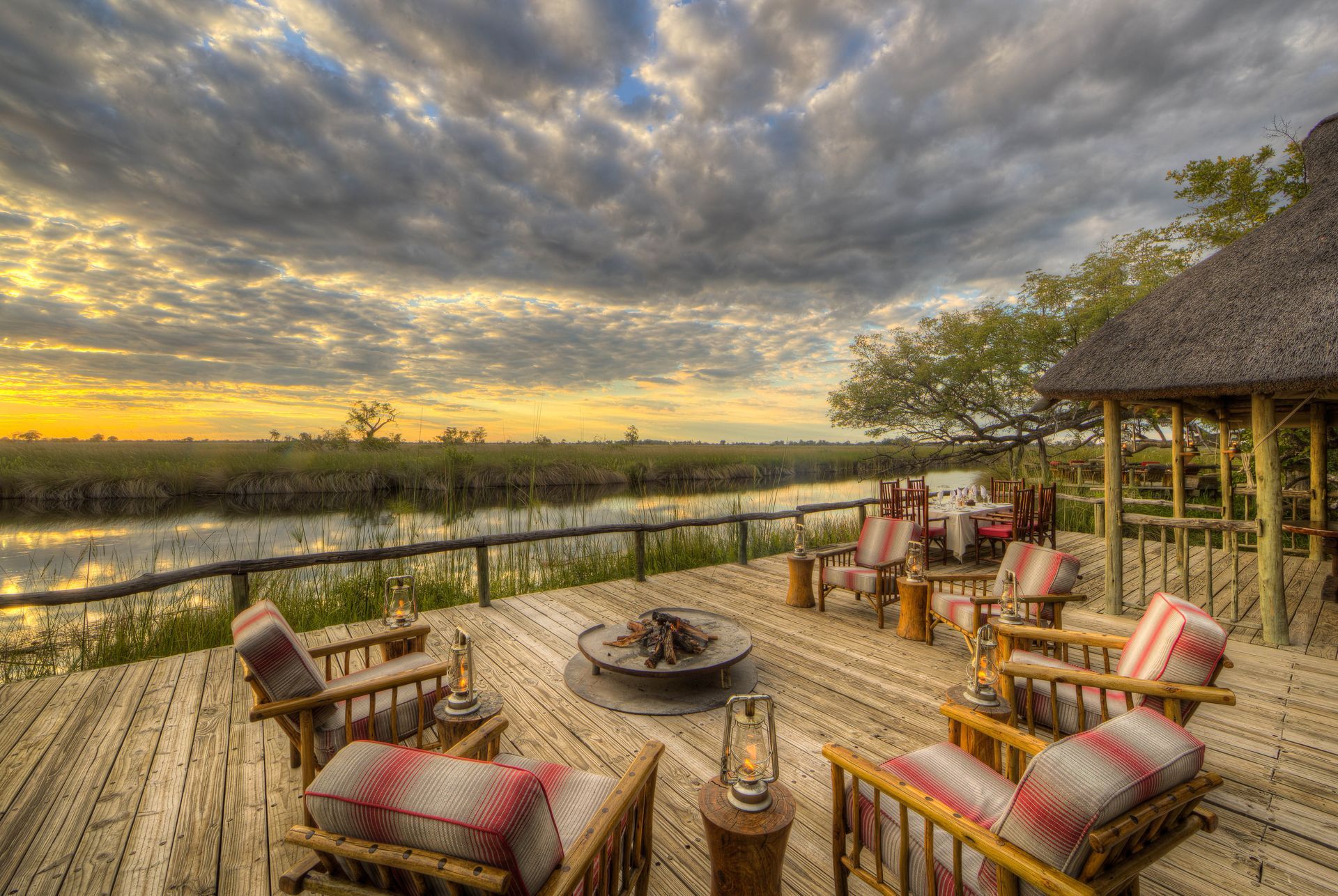 A scenic wooden deck overlooking a marsh at sunset, featuring comfortable seating arranged around a fire pit.