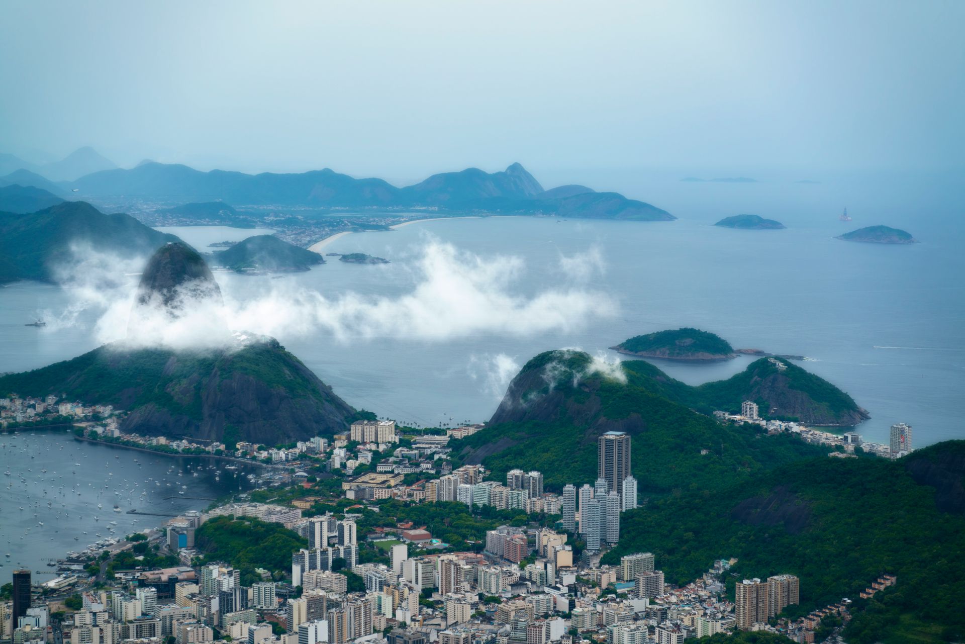 An aerial view of Rio de Janeiro featuring the iconic Sugarloaf Mountain partly shrouded in clouds above coastal cityscapes.