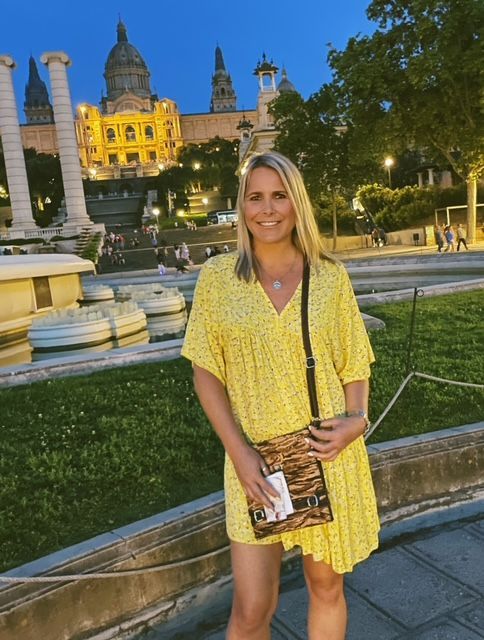 A smiling person in a yellow patterned dress stands before the illuminated Museu Nacional d'Art de Catalunya in Barcelona.