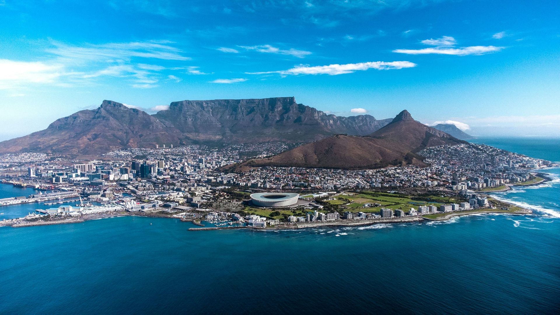 Aerial view of Cape Town, South Africa, featuring Table Mountain, the stadium, and the coastline against a blue sky.